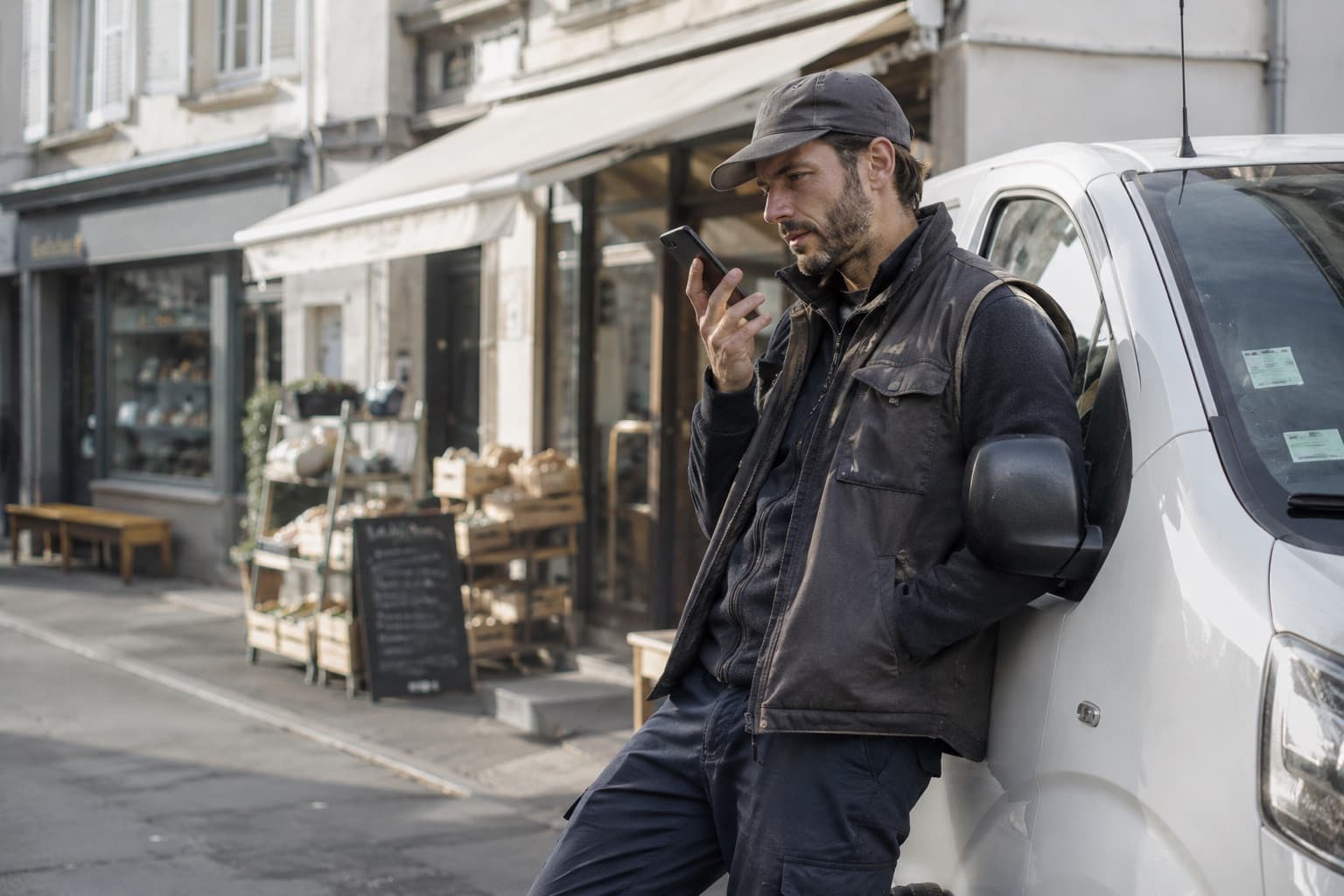 Un livreur adossé à son utilitaire dicte une note devant un commerce